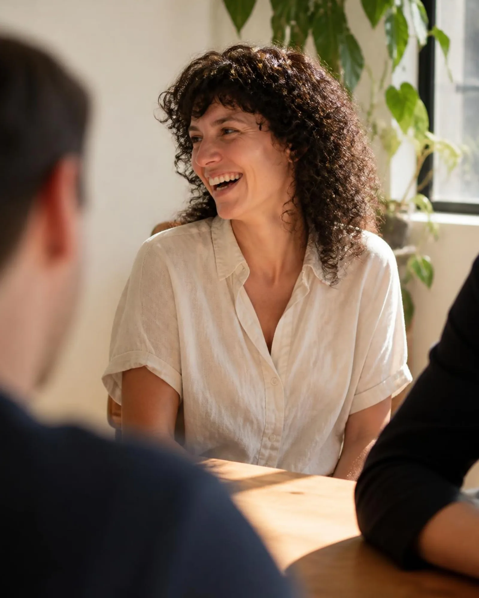 A woman laughing in a small team meeting by a sunny window.